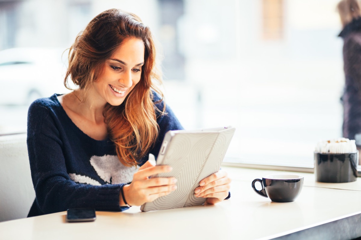 A woman smiling while reading a tablet at a cafe table, with a coffee cup beside her